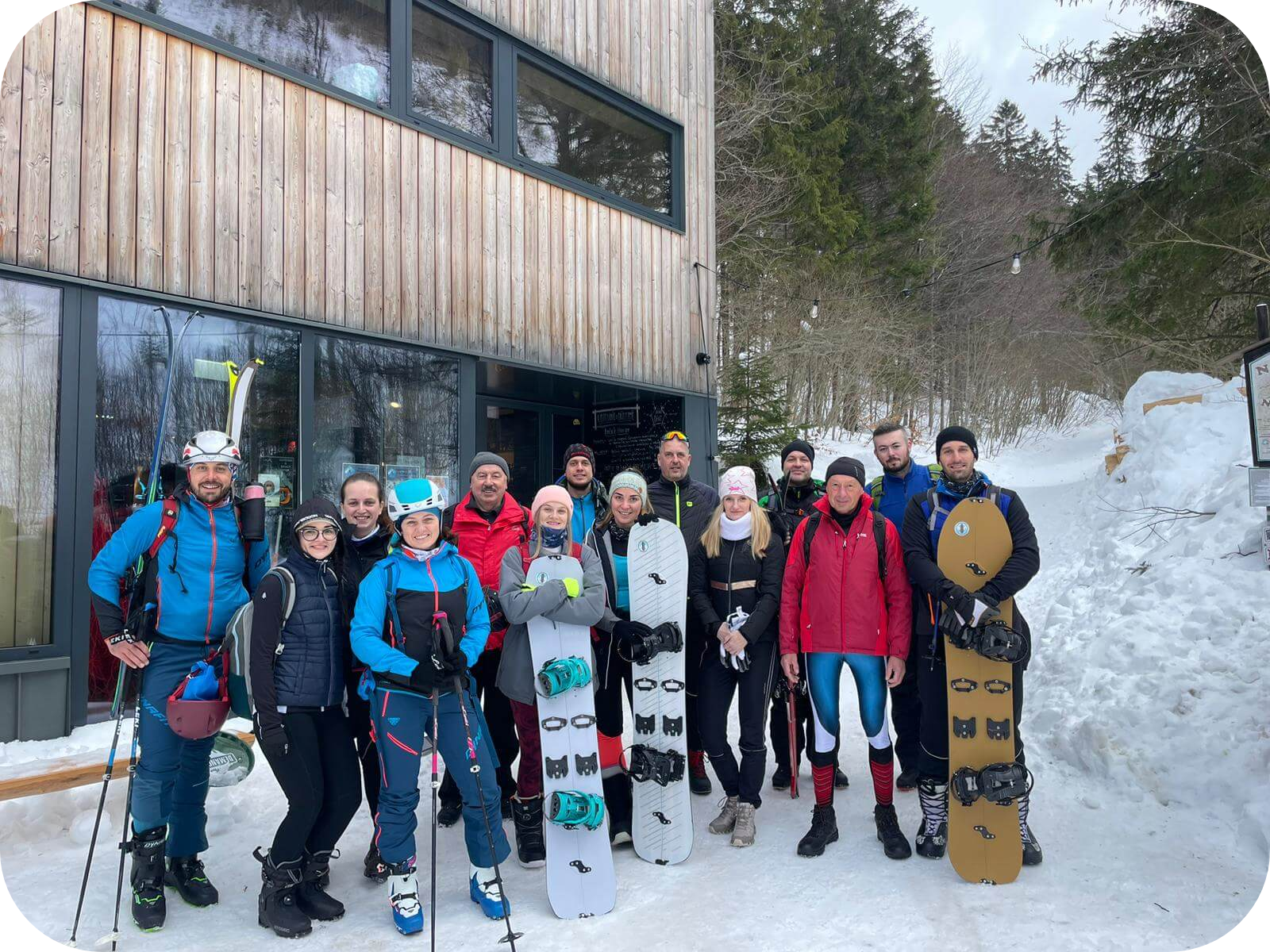 Zamestnanci spoločnosti JKBOZ stoja v zime pred chatou s lyžami a snowboardami, oblečení v teplom oblečení.